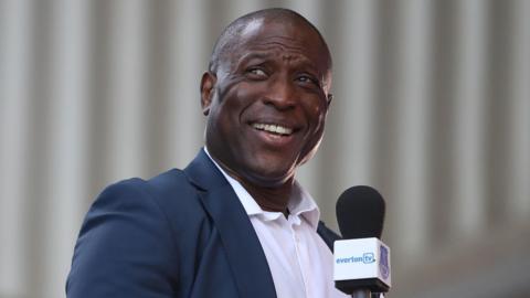 Kevin Campbell, wearing a dark navy suit jacket over a white shirt with an open collar, smiles while holding a microphone with the branding EvertonTV. 