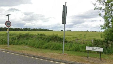 A view of a green field with a white sign reading Sandy Lane West.