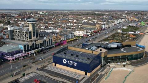 An aerial view of Great Yarmouth seafront. The view shows the Marina Centre, a large blue building with "Marina Centre" in large white letters one side. To the right is the beach, and the left shows a mixture of retail buildings, hotels and housing. 