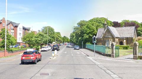  Albert Road near Hesketh Park. Three-storey blocks of modern-built flats line the left hand side of the road while a gatehouse stands at the entrance to Hesketh Park on the right side of the road.