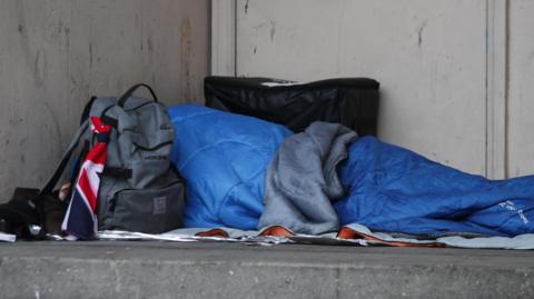 A homeless person in a blue sleeping bag sleeping in a doorway and surrounded by personal belongings including a backpack 