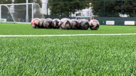 The image shows a close‑up view of an artificial 3G sports pitch, with the camera positioned low to the ground so the synthetic grass fills most of the foreground. In the background, slightly out of focus, there is a row of footballs lined up in a straight line near a white pitch marking. Further back goalposts can be seen, along with fencing and tall trees surrounding the pitch.
