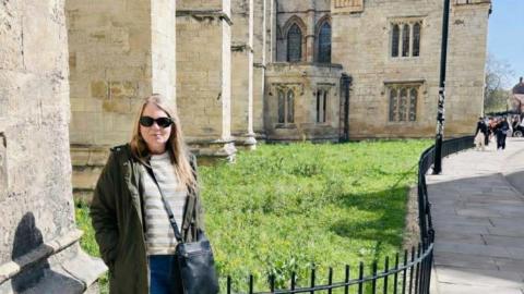 Gemma Louis with long blonde hair stands in front of the grassed area to the side of large building - a minster or cathedral. She is wearing sunglasses, a dark green anorak-type jacket over white and brown striped top, with a dark blue leather bag hung crossways over her shoulder.