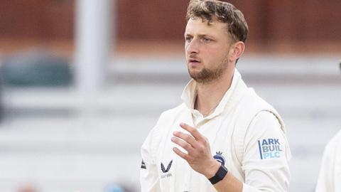 Luke Hollman dressed in cricket whites, in front of an umpire wearing a large hat, white jumper and black trousers
