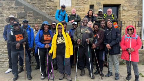 A group of twenty people, clearly walkers (many with walking poles, all wearing walking boots and raincoats) are gathered together. Several have T-shirts with the groups name 'We In Front' on them. They are stood in front of Castle Hill in Huddersfield, and are all looking directly at the camera smiling. They are aged between 50 and 80, with one younger member. 