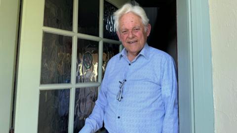 Pierre Tostevin standing in his doorway, he is wearing a blue shirt