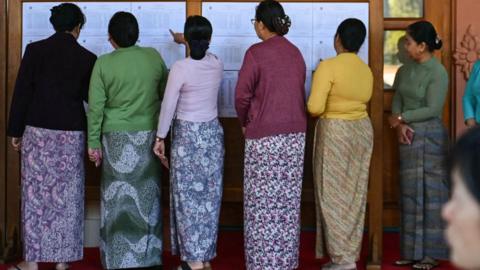 Women line up at a polling station in Naypyidaw on December 28. They are wearing cardigans over long, traditional outfits as they look at a notice board with their backs to the camera.