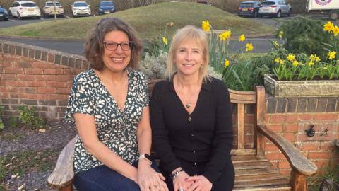 deborah and mel are sitting on a wooden bench in the garden at the shakespeare hospice. Mel is wearing a black dress and deborah is wearing a green and white patterned top with blue jeans