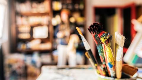 A pot of dried paintbrushes sitting on a table in the foreground. In the blurred background there is a woman standing in front of a wooden bookshelf full of art supplies. 