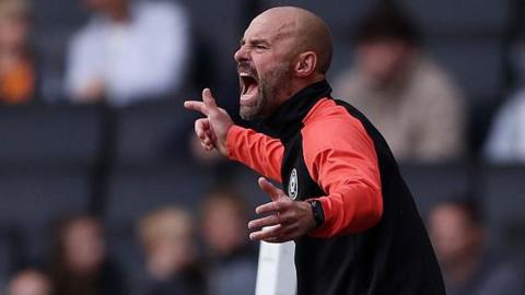 MK Dons boss Paul Warne in animated pose, shouting from the sidelines with his arms outstretched