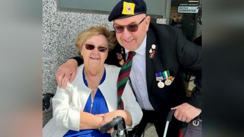 Roy and Marian Muir. Mrs Muir is sat down while Mr Muir has his arm around her and is crouching down. Mrs Muir is wearing a blue top with a white blazer, while Mr Muir is in a black suit, adorned with army medals. They both wear sunglasses.