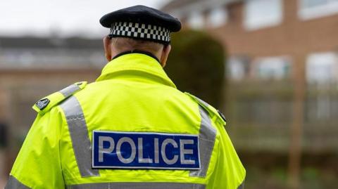 A police officer wearing a hi-vis jacket walking around what appears to be a housing estate.
