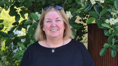 A woman in a navy top stands in front of a garden fence smiling at the camera. 