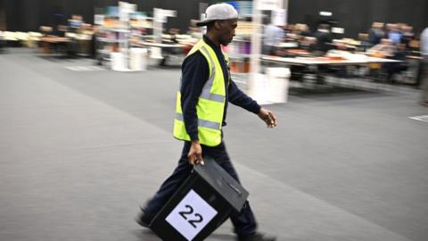 A man in a hi-vis and baseball cap worn backwards carries a ballot box with the number 22 on it. The background in the hall is out of focus.