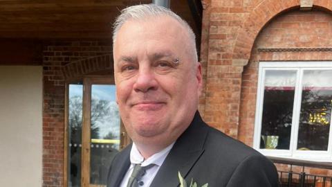 Sean is short cropped grey hair and a ring piercing on the end of the right eyebrow. He is wearing a black blazer over a white shirt and grey tie. There is a flower in the right breast pocket. He is standing in front of a redbrick building and smiling into the camera.