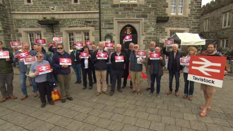 Group of campaigners holding up rail signs saying Tavistock, showing support for the reinstatement of the Tavistock to Plymouth Rail link.