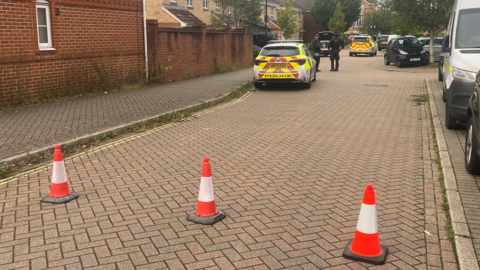 Orange cones block off a residential road. Two police officers stand next to a police car beyond the cones. There are other emergency vehicles in the distance