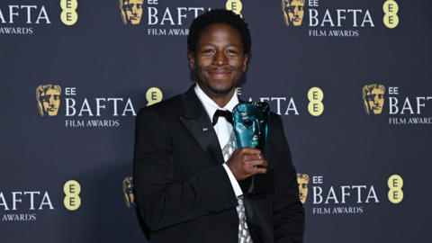 David Jonsson stands in front of a hoarding with "EE Bafta Film Awards" printed on it. He's wearing a black suit with white shirt and bow tie.