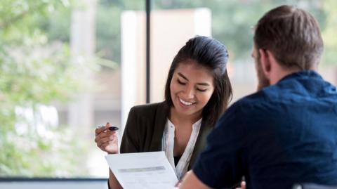 A woman is smiling and looking down at a piece of paper with a pen in her other hand. She is wearing a blazer and shirt. Opposite her is the back of a man in a blue shirt. Behind them is a large window