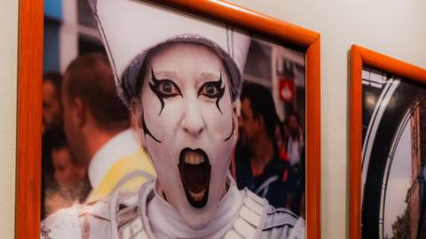 A picture of a person wearing white and black face paint with his mouth wide open. He is wearing a white and silver costume and there is a crowd of people passing behind him. The picture is mounted in a brown/orange frame on a white wall.