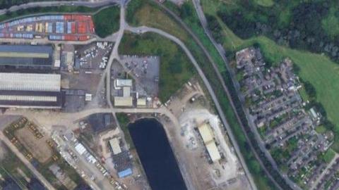 A satellite view of part of Port Clarence with the site where the proposed hydrogen production plant may be built in the middle. A housing estate is shown on the right of the picture. From there, and moving to the left, there is a road, then a thin line of trees followed by a single track railway line. There is further greenery before a road on the edge of the industrial site. That has a few buildings dotted around a wharf on the River Tees, the water dominating the bottom centre of the image. Much of the land around it is empty and used for parking, but there is a huge industrial unit and lots of shipping containers in the top left.