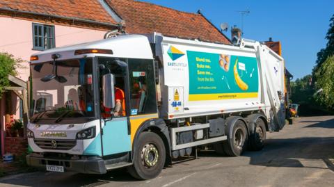 A waste collecting van with council staff sitting inside it wearing high-vis orange clothing. The van is white with blue and yellow council branding on it. There are cottages behind it. It is a sunny day with a blue sky above.