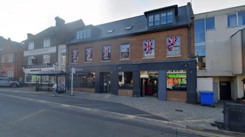 A Google Maps image of Knox bar in Dorking. There is a black lower part to the building and brick higher up. There are a number of Union Jack flags on the building. 