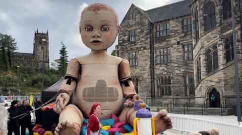 A large puppet of a unclothed baby sits on top of a pile of pink, blue and yellow custions. The puppet - around four times the height of adults stood nearby - has short ginger hair and blue eyes, and is holding a purple and yellow dummy in one hand. A giant purple and yellow bottle, made to look like it is filled with milk, stands at its feet.