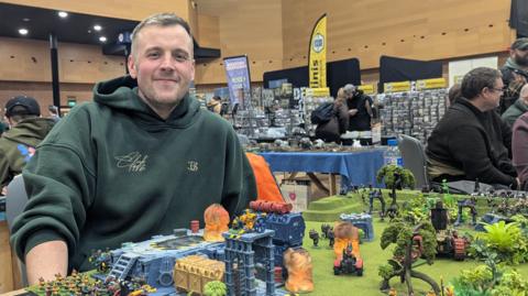Alex Nicholson, a man with fair hair wearing a green hoodie, sits in front of a board covered in miniature Warhammer figures, structures and trees. 