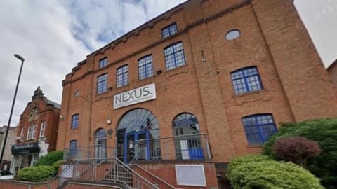 A brown building which has glass windows with dark blue frames. It has a sign which says "Nexus" in capital letters and underneath on the same sign it says: "Institute of Creative Arts" 
