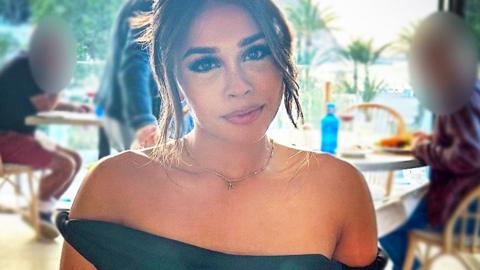 Ravika sitting in a restaurant, pictured head and shoulders looking directly at the camera. She has her hair tied up and is wearing a black off-the-shoulder dress.