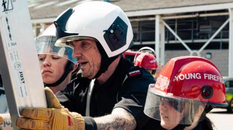 A close-up of firefighter in his uniform and helmet. He is holding a ladder. There is a school pupil either side of him. They are also wearing fire helmets.