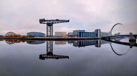 Bridges, buildings and a large decommissioned crane are reflected in the still waters of the River Clyde