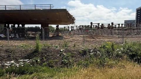 The edge of the flyover, which is being demolished bit by bit, revealing the jagged edge of the thick, concrete structure, suspending in the air. Steel posts stick out in all directions, like the end of a frayed rope, which run through the concrete to reinforce it. Beneath the bridge is earthy ground, surrounded by a chainlink fence.