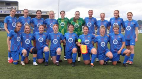 A team of female footballers pose together in two rows on a green pitch wearing a blue kit. The front row is on one knee, the back row is standing, and there ate two goalkeepers in the centre of the back row wearing a green kit.