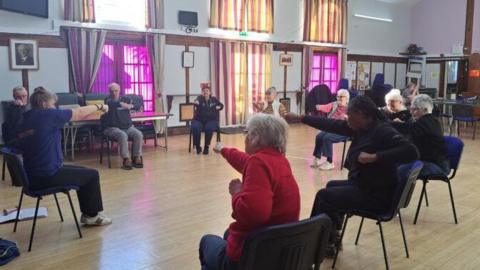 Ten people sit in a circle for a chair exercise session in a large hall with a wooden floor and tall windows. They are extending their arms in a punching pose. 