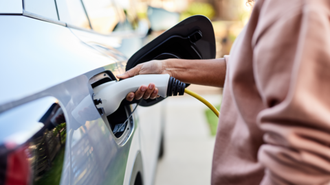 A person in a pink jumper holding a charging cable to the side of a silver car