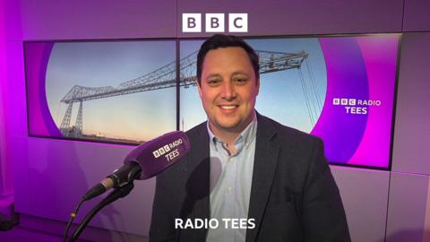 Mayor Ben Houchen smiling in the BBC Radio Tees studio. He wears a dark suit jacket and light blue button up shirt and sits in front of a large microphone. Behind him is a screen depicting the Transporter Bridge and the BBC Radio Tees logo. The studio lights give it a purple hue.