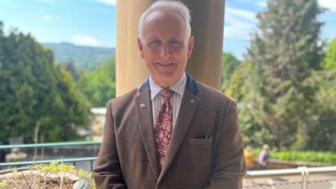 Man with short grey hair, brown tweed jacket stands on balcony with hillside backdrop