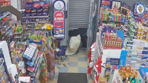 CCTV picture of a shop full of stock, a man is hunch over by the shutters covering the front door preventing him from escaping. He's holding a bag containing stolen goods. You can also see products that have been displaced on the floor.