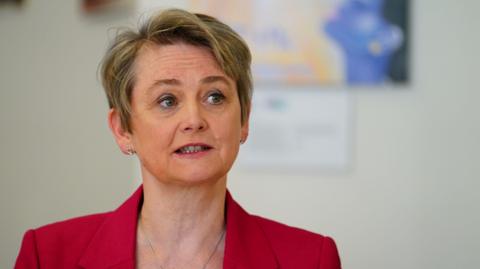 Yvette Cooper pictured in profile from the shoulders up - she has short fair hair and is looking off to her left. She is wearing a red jacket. 