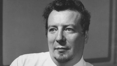 Stuart Burrows, shown in a black and white image, looks away from the camera wearing a shirt and tie. He has short dark hair and a goatee.