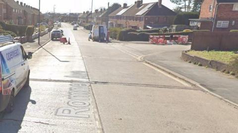 A suburban street with red brick houses. There are road traffic barriers for roadworks to the right of the screen, and two BT Openreach vans parked up. 