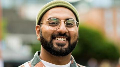 Akshay Sharma looking at the camera wearing round glasses and a green hat.
