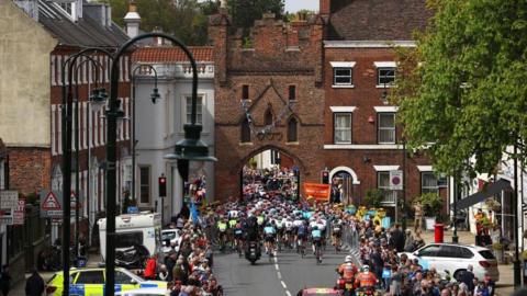 A large group of cyclists followed by a motorbike race through Beverley's North Bar - an ancient red brick archway. Crowds of people stand at barriers at the roadside.