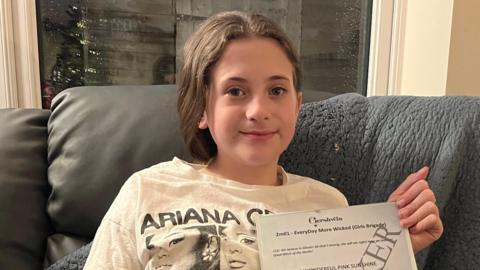 Lily sits on a grey sofa. She is wearing a t-shirt with Arianna Grande's face on it and she holds a framed piece of a paper