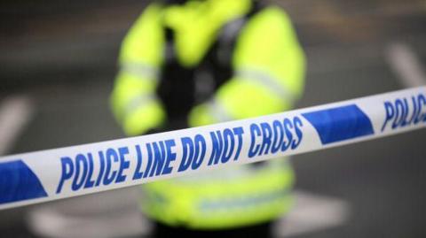 Police tape in blue and white stretches across the frame, a police officer in hi vis jacket and black body armour is standing behind the tape. 