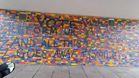 A brick wall in a tunnel. Around 1,500 bricks are individually painted bright colours, some with symbols and flags, others with animals and local landmarks. Over the top of it someone has spray painted the phrase "Vote Reform UK and this all goes away" 