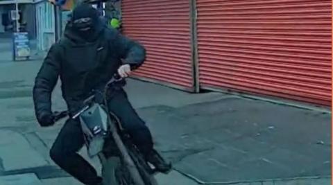 E-motorbike rider, wearing all black clothes and a balaclave, mounts the pavement