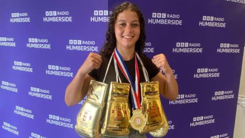 A young girl with dark brown curly hair is smiling at the camera and holding up both her fists. She is wearing a black top and around her neck are three gold boxing gloves and a gold medal with blue, white and red stripes on the ribbon. In the background is a purple wall which repeatedly says 'BBC Radio Humberside'.